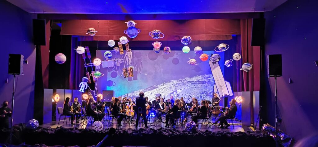 występ na scenie, na pierwszym planie tyłem stoi dyrygent, za nim orkiestra , nad glowami wisza kolorowe planety dekoracje są w stylu kosmicznym 
