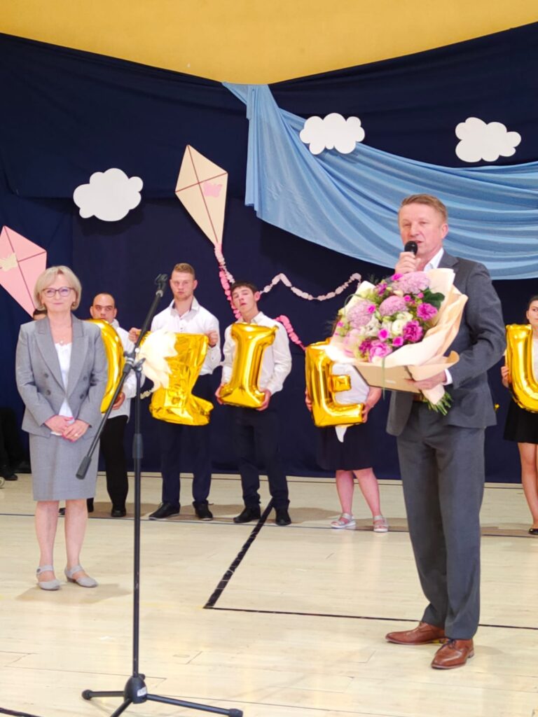 Na sali gimnastycznej. Po lewej kobieta w eleganckiej szarej garsonce, po prawej przed mikrofonem mężczyzna w garniturze z wielkim bukietem różowych kwiatów. Z tyłu w białych koszulach dzieci trzymają przed sobą złote balony litery Z I Eny