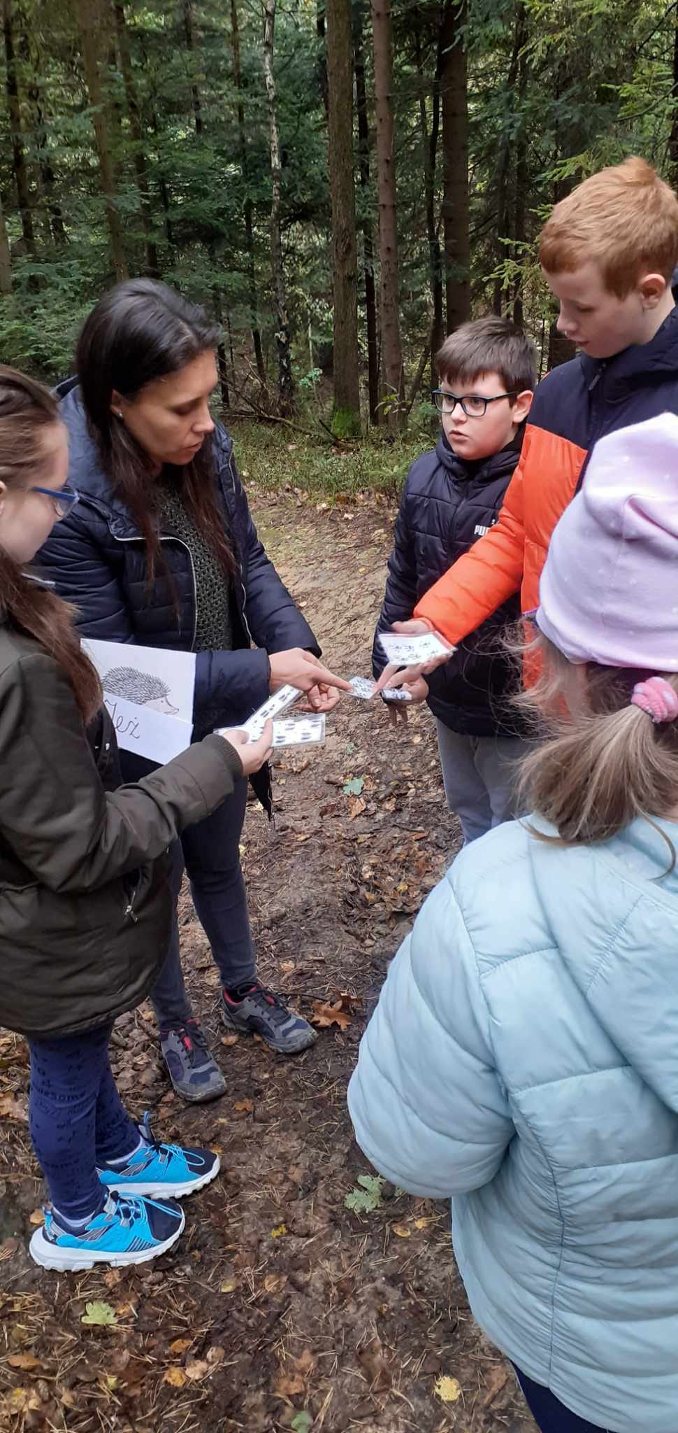 Nauczycielka wraz z uczniami są w lesie. Nauczycielka pokazuje uczniom karty pracy.