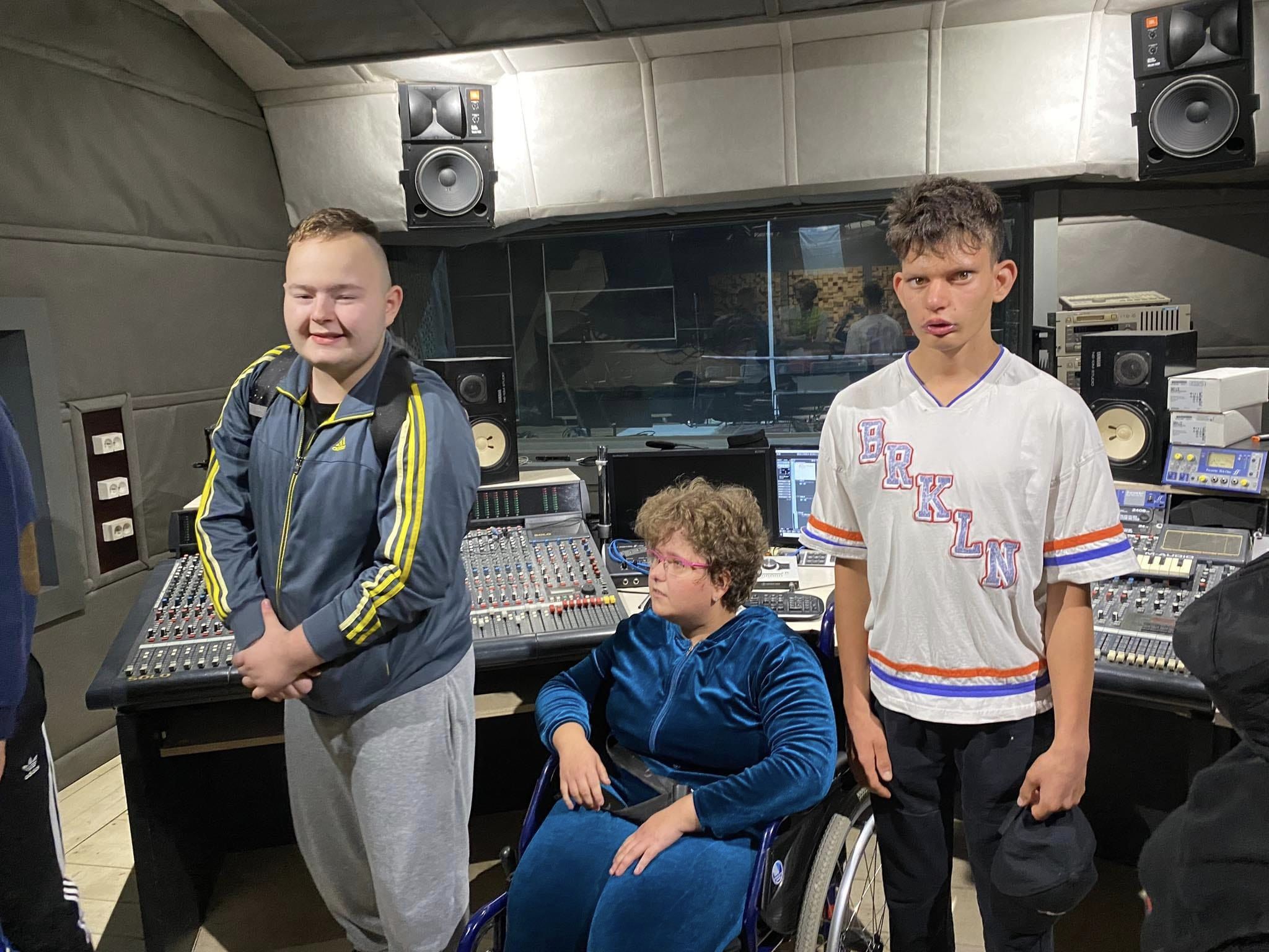 Na zdjęciu znajduje się dwóch chłopców stojących pomiędzy dziewczyną siedzącą na wózku inwalidzkim. Uczniowie znajdują się w pomieszczeniu realizatora dźwięku.