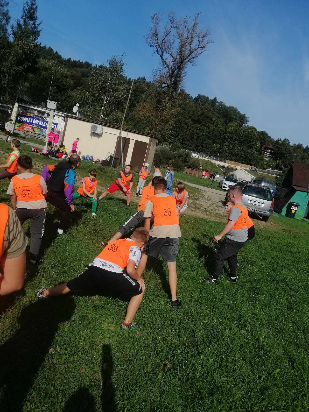Na zdjęciu są uczniowie ubrani w pomarańczowe koszulki. Uczniowie wraz z nauczycielem wykonują rozgrzewkę na trawniku.