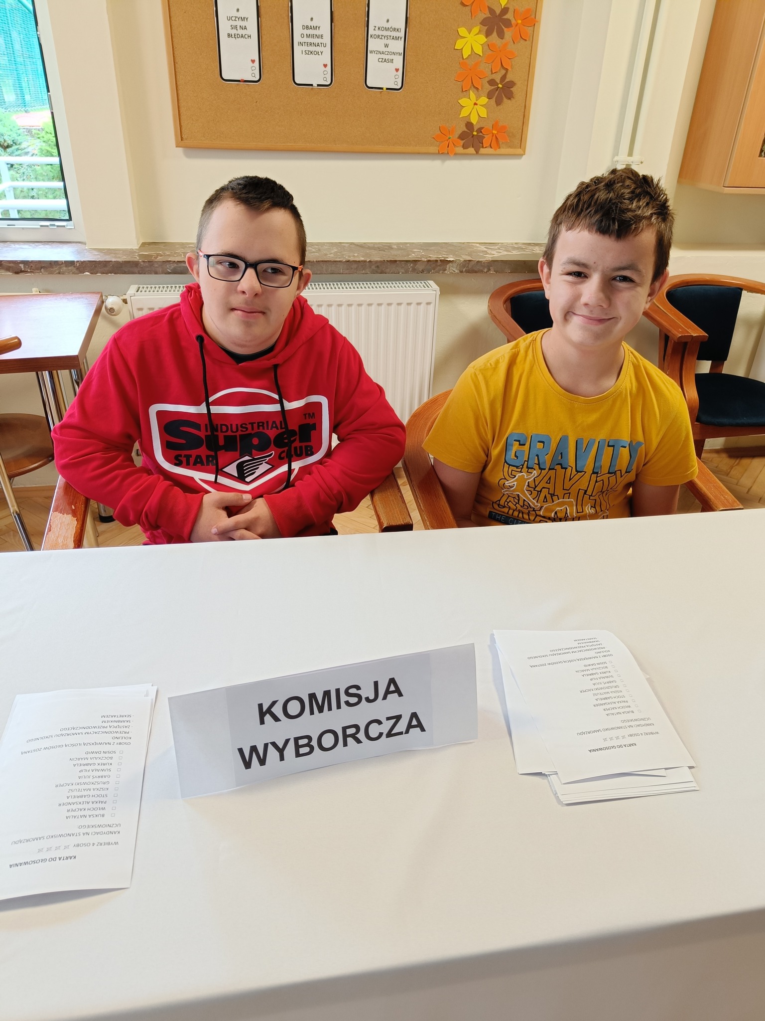 Na zdjęciu znajduje się dwóch chłopców siedzących przy stole. Na stole widnieje napis komisja wyborcza.