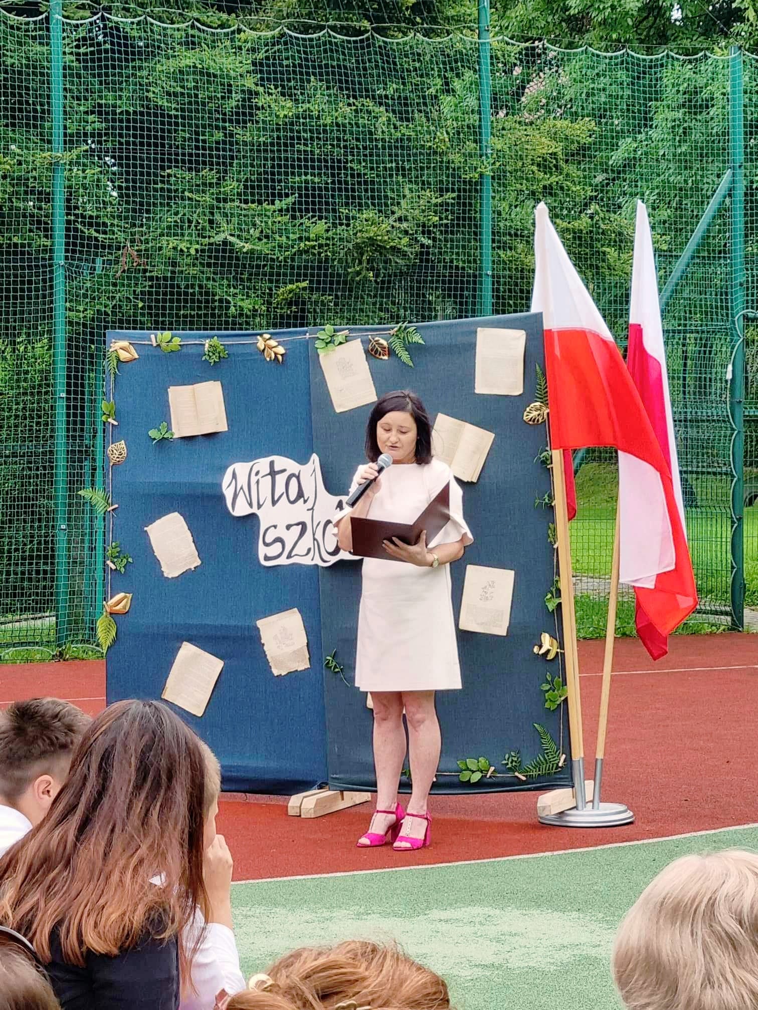 Na boisku szkolnym stoi kobieta w ręku trzyma mikrofon , przemawia. w tle  białoczerwone flagi i napis Witaj szkoło