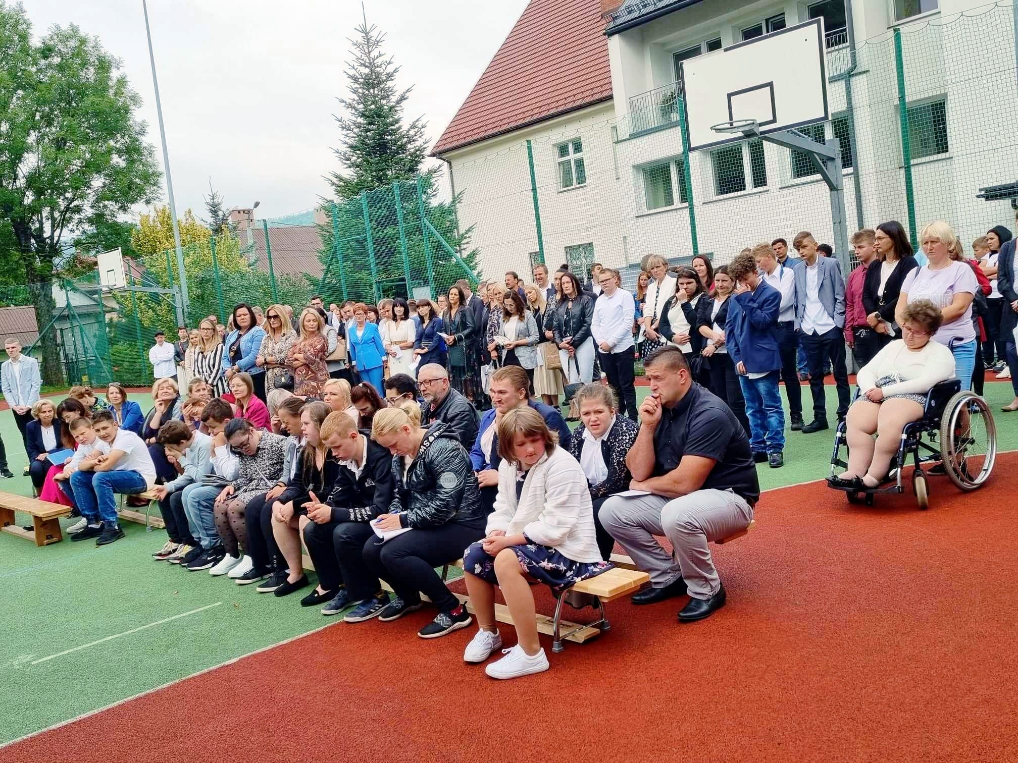 na boisku szkolnym na ławkach siedzą uczniowie i rodzice, z tyłu stoją uczniowie i rodzice