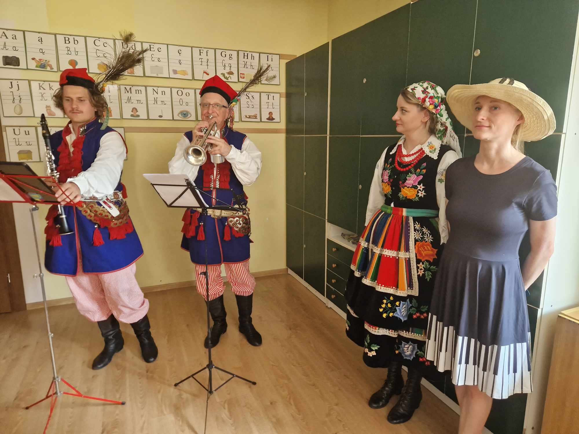 po prawej kobieta w sukience i kapeluszu, obok niej kobieta ubrana w strój regionalny krakowski, obok dwóch mężczyzn , jeden gra na trąbkach