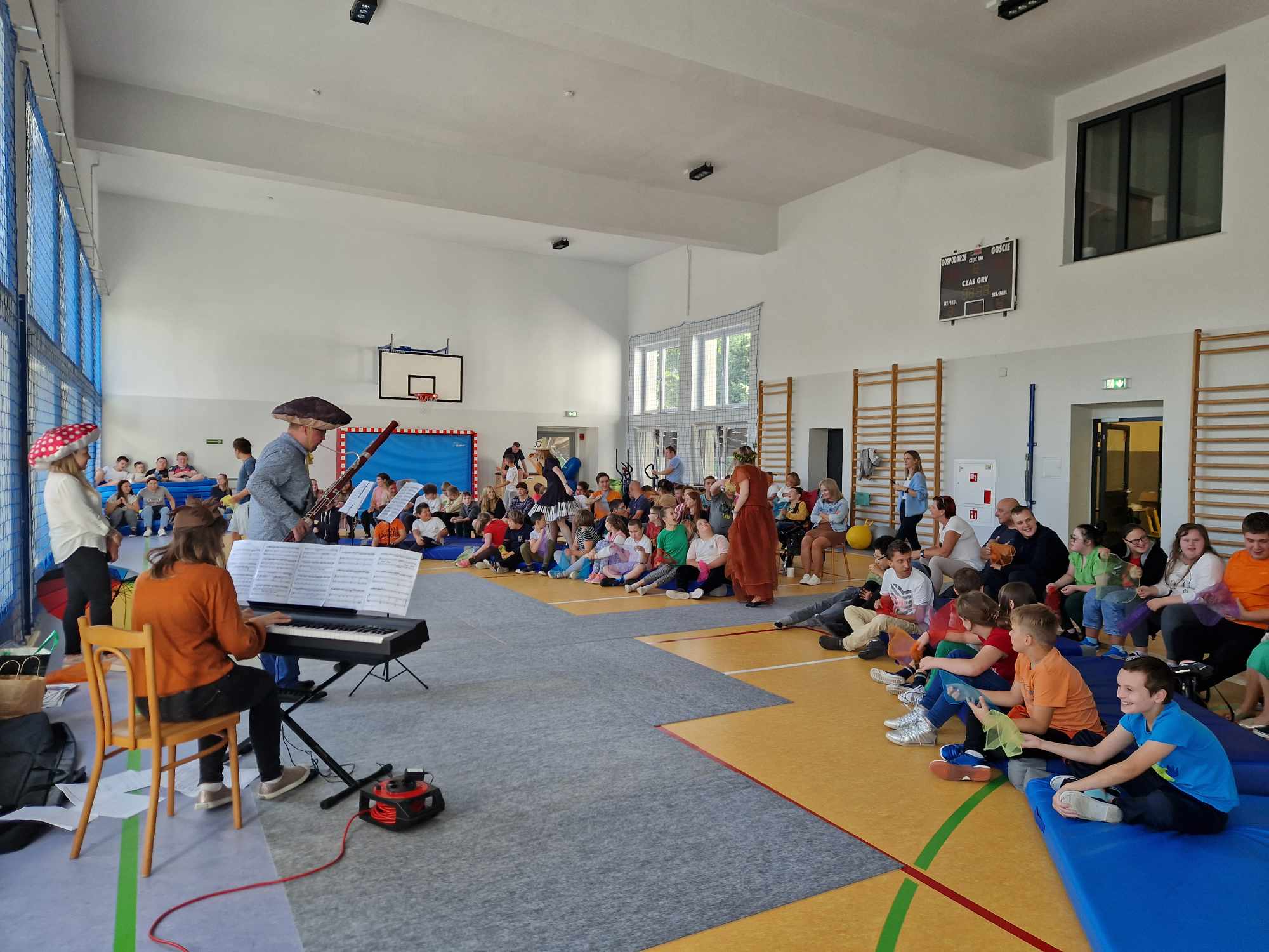 Na zdjęciu znajduje się duża grupa dzieci siedząca po turecku na sali gimnastycznej. Na przeciwko dzieci znajduje się pan, który gra na instrumencie zwany fagotem, a obok niego pani grająca na pianinie. 