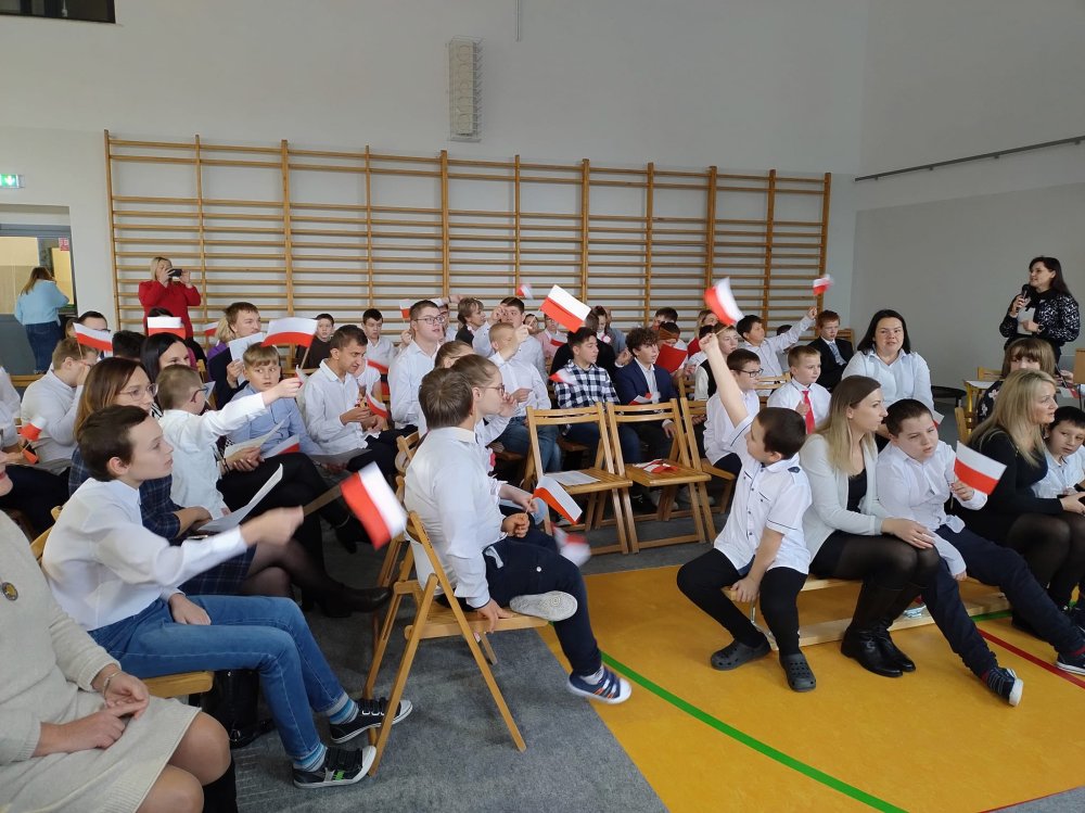 Na zdjęciu znajdują się uczniowie siedzący na krzesłach na sali gimnastycznej i wymachujący flagą Polski.