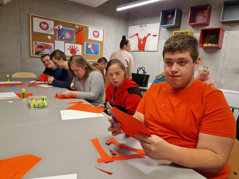 Na zdjęciu znajdują się uczniowie siedzący przy stole, którzy wycinają wzór kotylionu z czerwonej i białej kartki.
