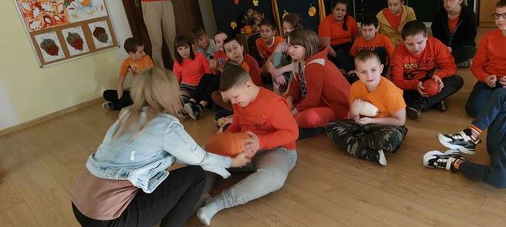Zdjęcie przedstawia uczniów ubranych w pomarańczowe koszulki siedzących na podłodze oraz nauczycielkę wręczającą dynię uczniowi.