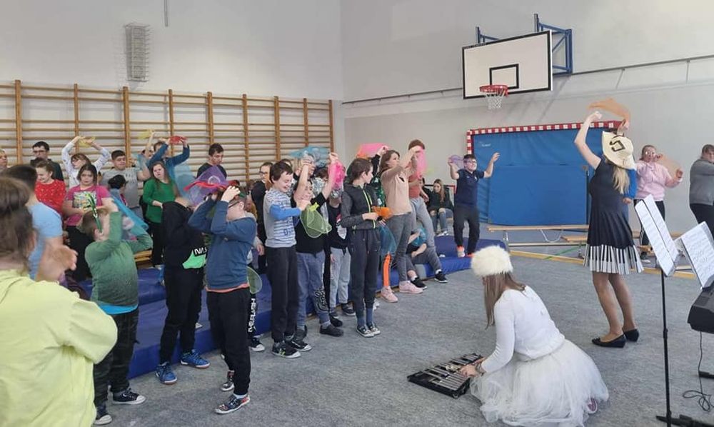 Na zdjęciu uczniowie stoją zwróceni twarzami do Pani Śnieżynki grającej na cymbałkach. Uczniowie pokazują ruchy do piosenki.