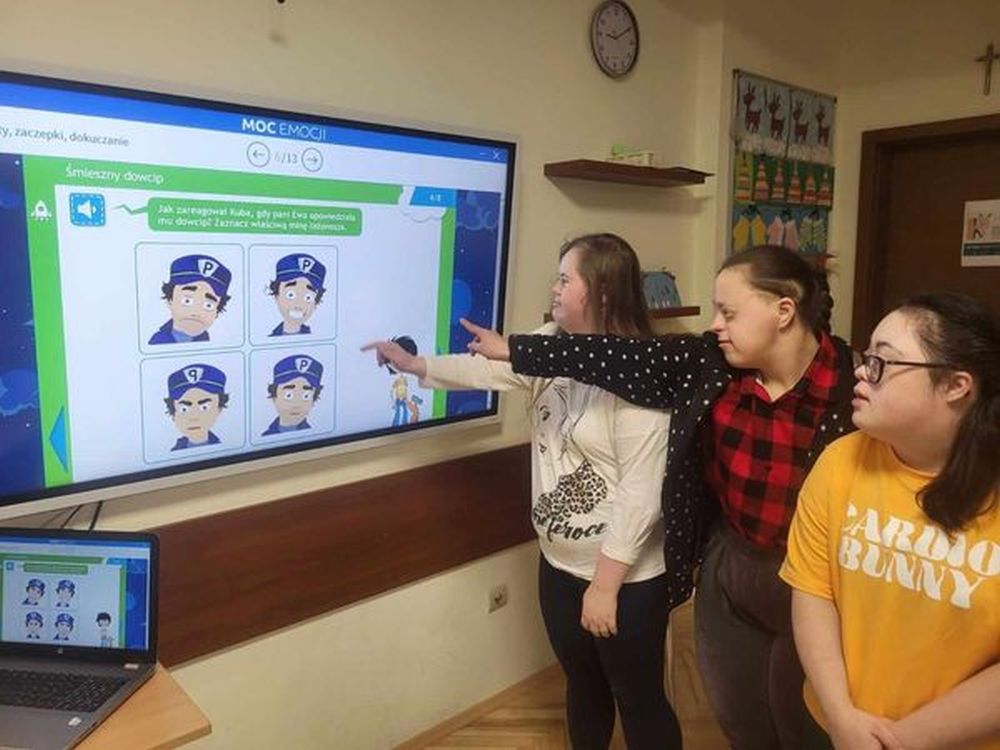 Zdjęcie przedstawia uczniów stojących przy tablicy interaktywnej. Uczniowie dobierają odpowiednie obrazki do sytuacji.
