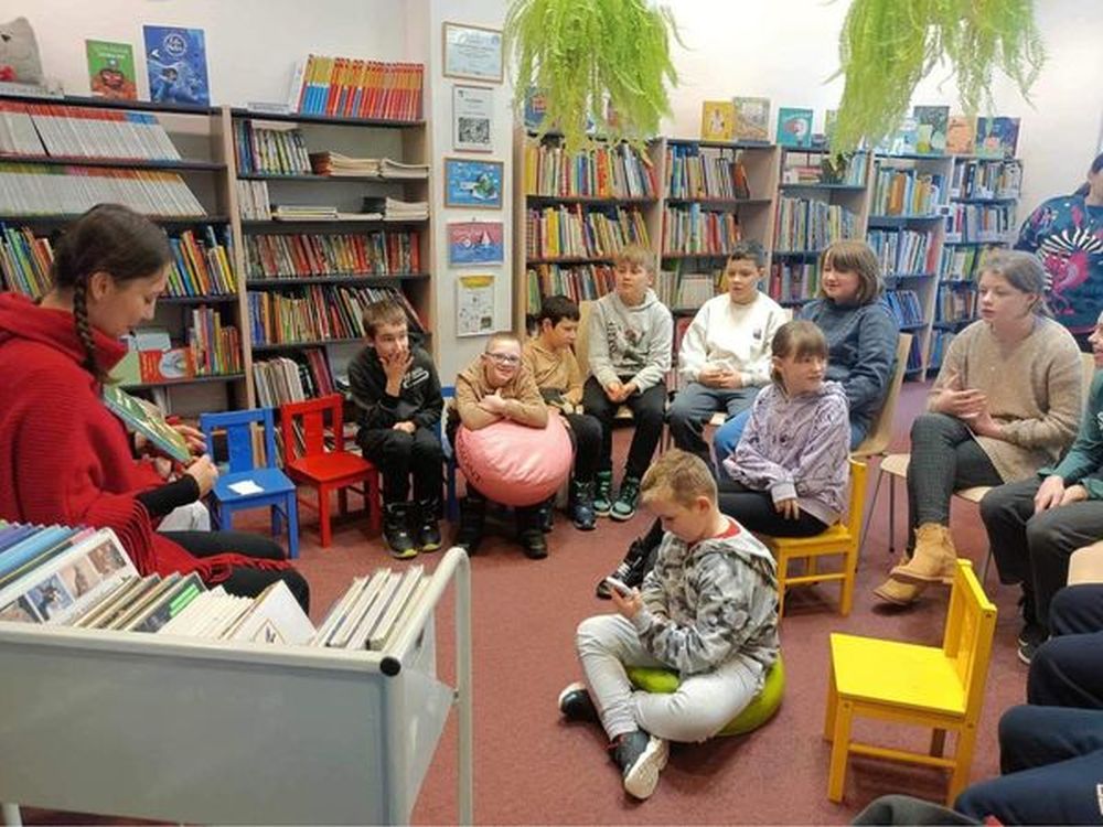Zdjęcie przedstawia uczniów siedzących na krzesełkach w bibliotece oraz panią bibliotekarkę pokazującą dzieciom książkę.