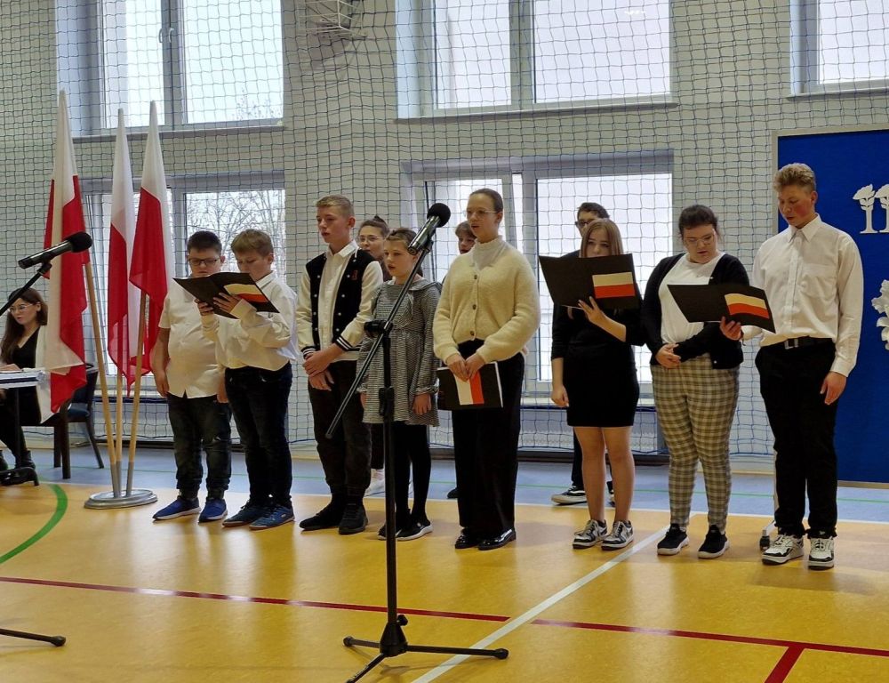 Grupa uczniów ubranych w białe koszule i czarne spodnie i spódnice stoi w rzędzie. Obok nich z lewej strony flaga Polski