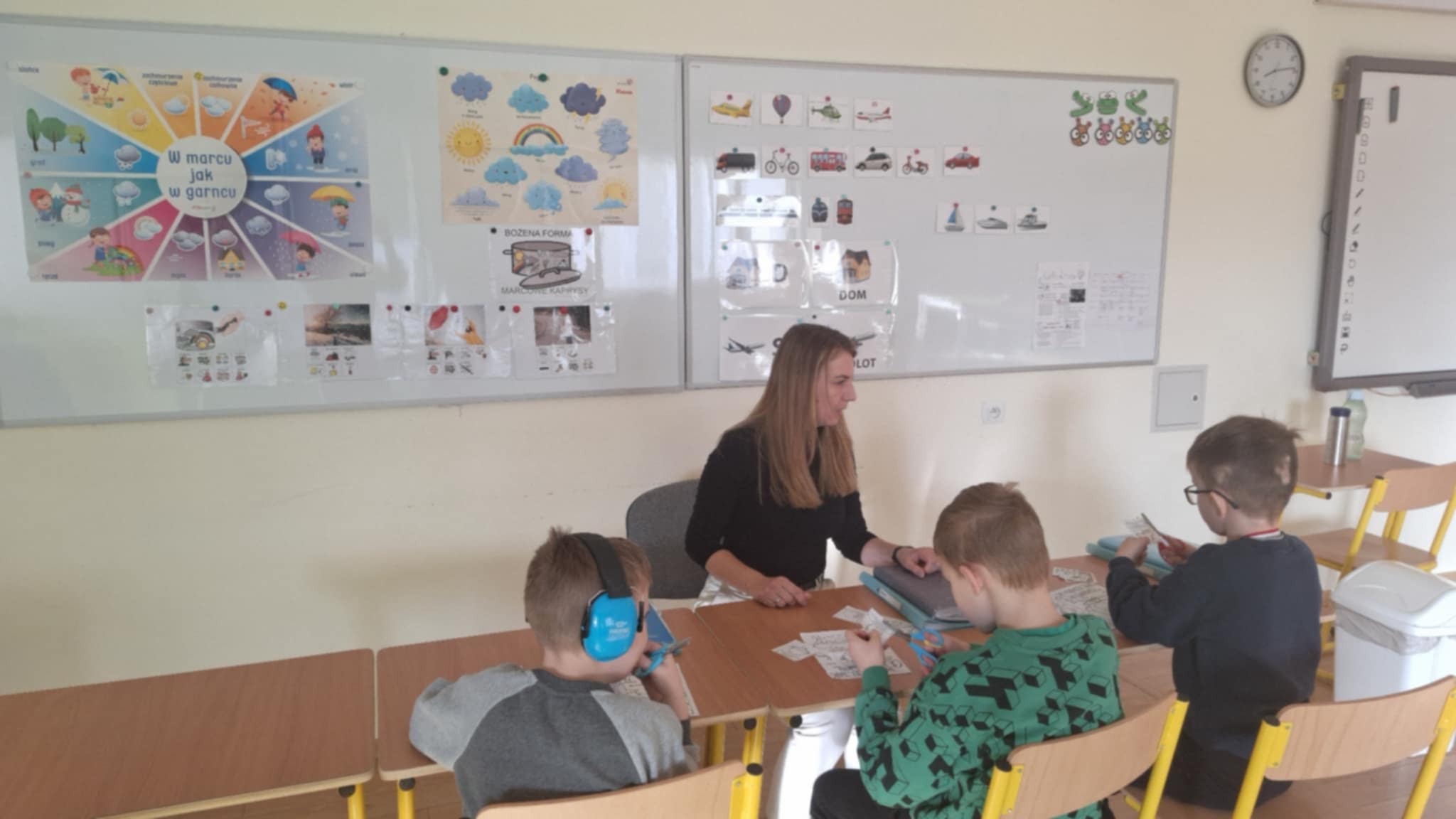 Nauczycielka siedzi zwrócona do uczniów, którzy siedzą w ławkach. Za nimi 2 białe tablice .