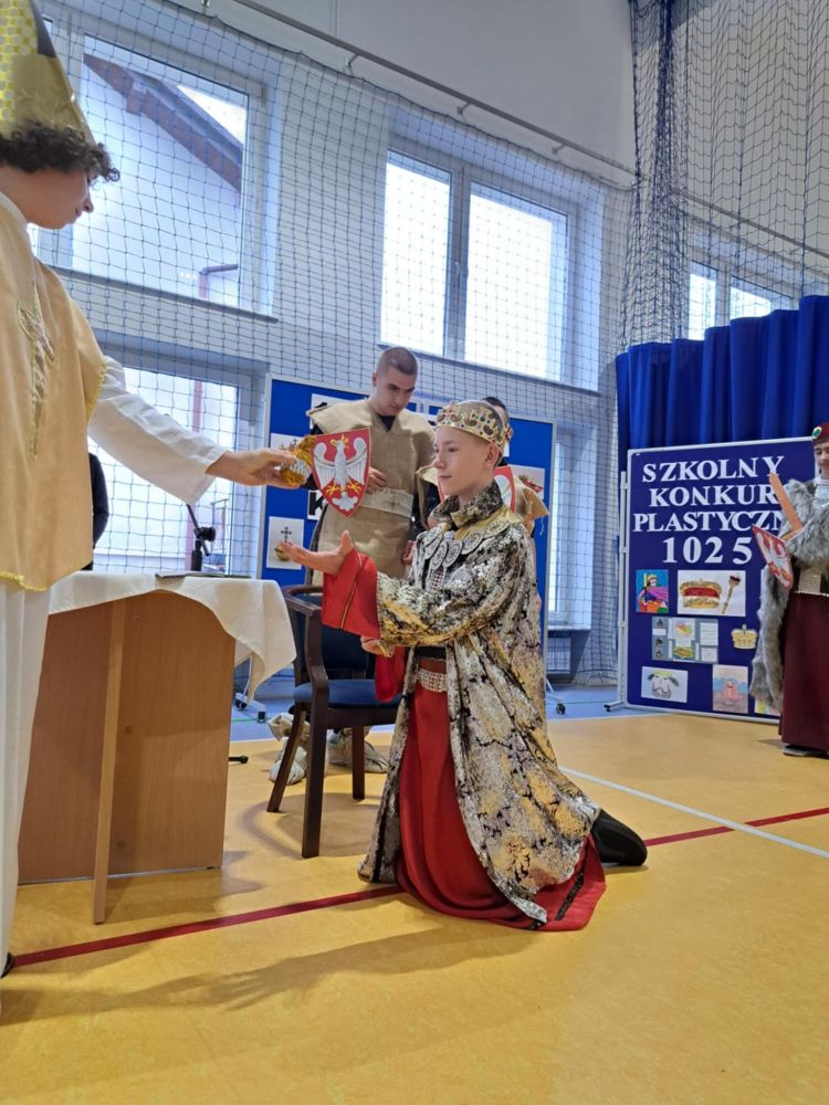 Uczniowie podczas przedstawienia z okazji 1000-lecia Korony Królestwa Polskiego. Zdjęcie przedstawia klęczącego Króla, za nim napis Szkolny konkurs plastyczny 1025.