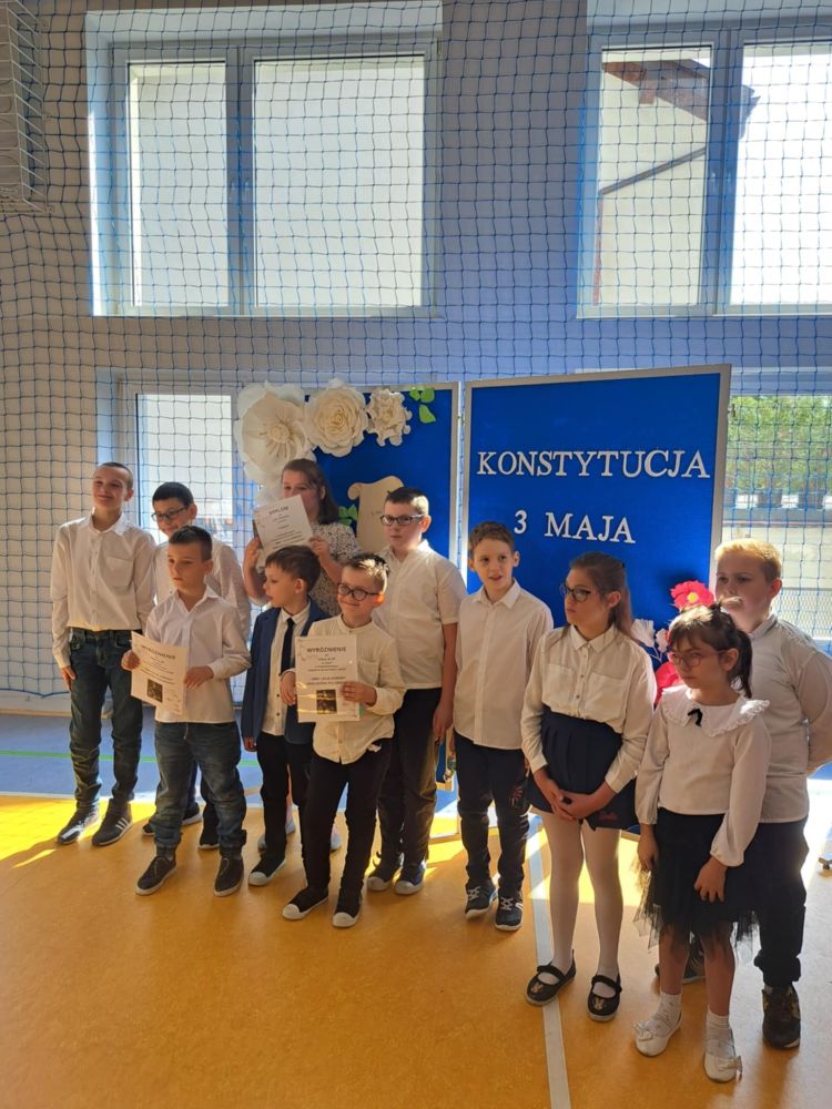 Grupa uczniów ubrana na galowo stoi na sali gimnastycznej, za nimi napis Konstytuacja 3 Maja na granatowym tle.