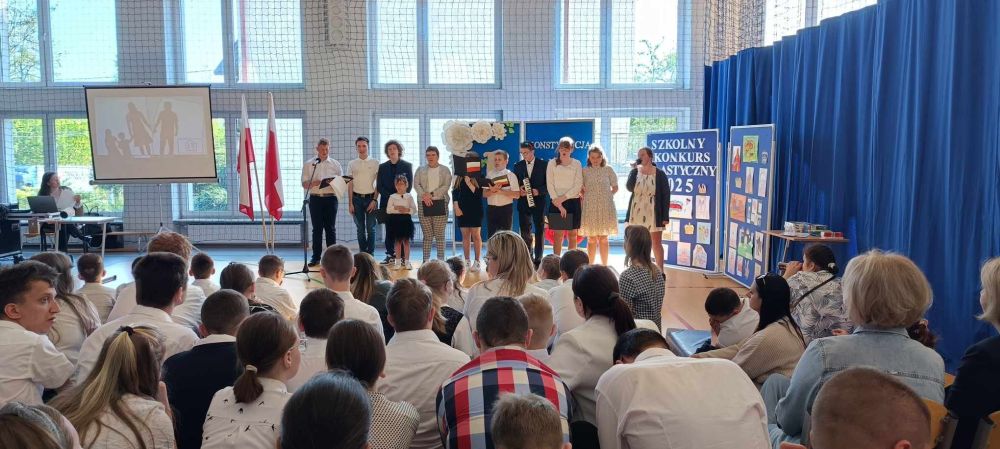 Grupa uczniów ubrana na galowo stoi na środku sali gimnastycznej podczas akademii. Przed nimi siedzą uczniowie i nauczyciele. Po lewej stronie stoją 2 flagi Polski.