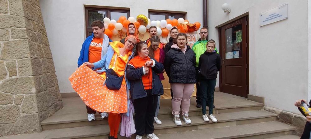 Grupa uczniów ubrana na pomarańczowo stoi na schodkach. Za nimi girlanda z białych i pomarańczowych balonów.