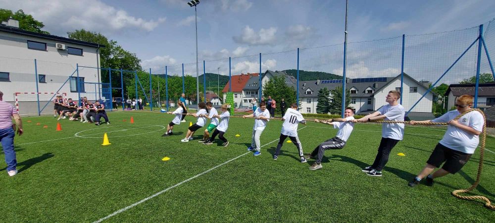 Grupa uczniów przebrana w białe stroje sportowe przeciąga linę na boisku sportowym.