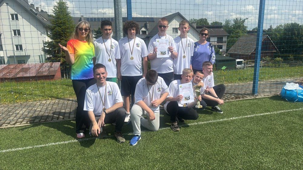 Grupa uczniów przebrana w białe stroje sportowe wraz z nauczycielami wf pozują do zdjęcia na boisku.