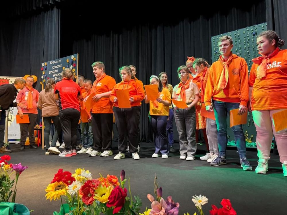Grupa uczniów Ośrodka ubrana na pomarańczowo stoi na scenie i śpiewa.