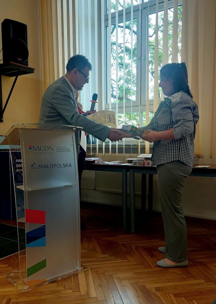 Uczennica odbiera dyplom z rąk pana ubranego w garnitur. Pan trzyma w ręce mikrofon i ściska dłoń dziewczynki.