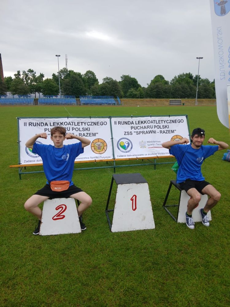 2 uczniów ubranych w granatowe stroje sportowe siedzą na podium, jeden z nich na 2 miejscu, drugi na 3 miejscu. Oboje mają podniesione ręce do góry ukazując ich siłę.