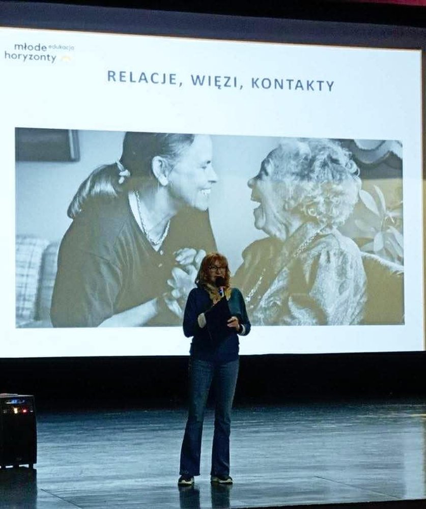 Pani w blond włosach mówi do mikrofonu, za nią ekran, na którym są 2 kobiety.