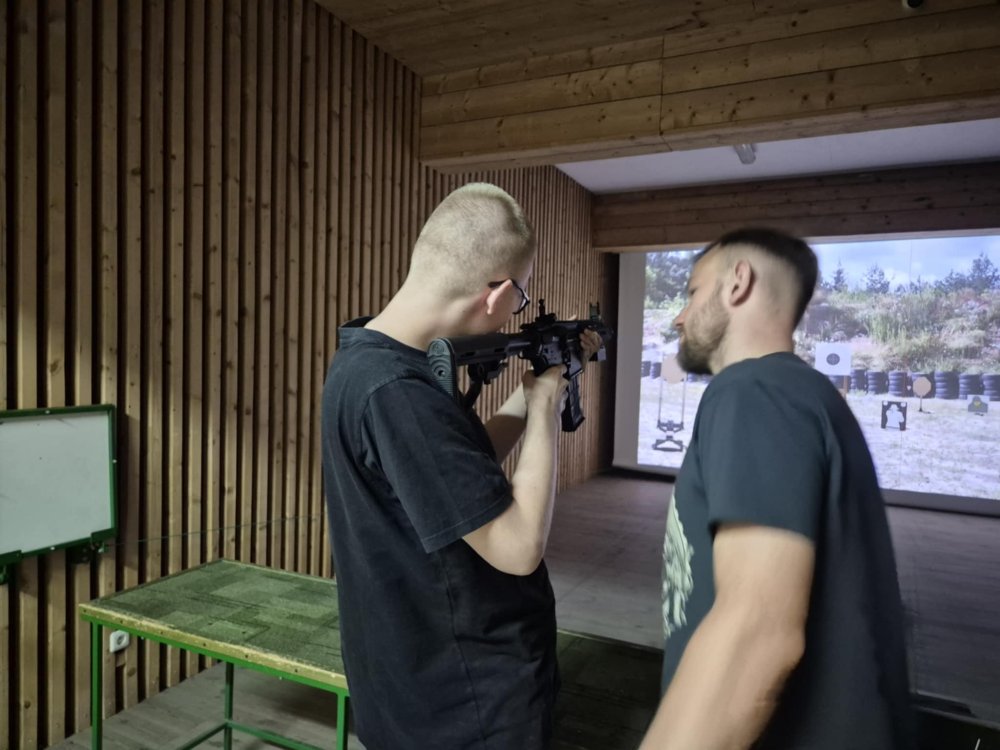 Uczeń trzyma w ręce strzelbę i celuje. Obok niego stoi pan, który pokazuje mu jak to zrobić.